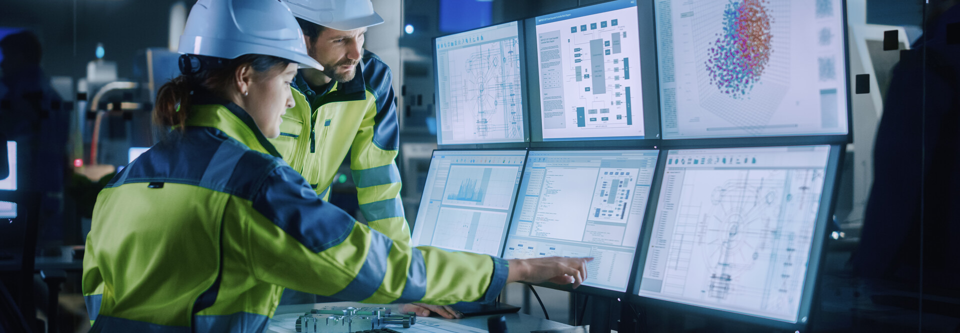 Two factory workers point to computer screens.