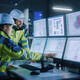 Two factory workers point to computer screens.
