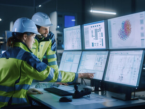 Two factory workers point to computer screens.